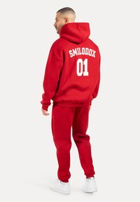 Conjunto de sudadera y pantalones de chándal de algodón rojo, con el texto blanco "SMILODOX 01" en la parte posterior, ajuste relajado y puños acanalados. Zapatillas blancas.