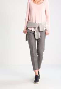 Helles rosa Langarmshirt, grauer Strickpullover an der Taille gebunden, graue maßgeschneiderte Hose und schwarze Sneaker mit weißen Sohlen.