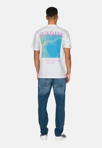 White cotton t-shirt with a large blue and pink graphic on the back featuring "SUNDAYS" text, paired with blue denim jeans and white shoes.