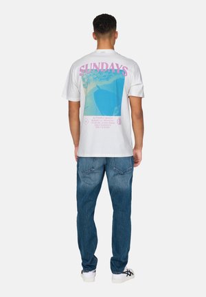 White cotton t-shirt with a large blue and pink graphic on the back featuring "SUNDAYS" text, paired with blue denim jeans and white shoes.