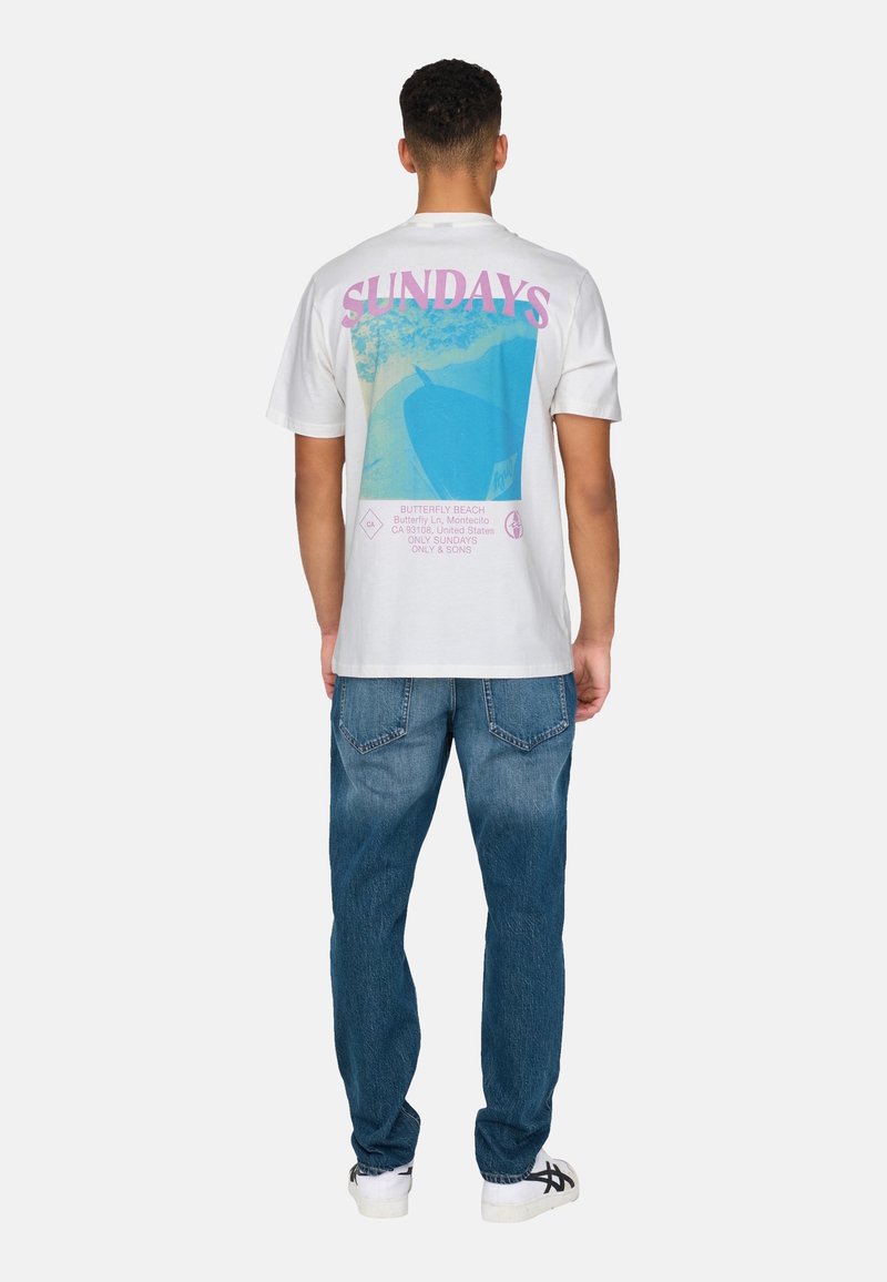 White cotton t-shirt with a large blue and pink graphic on the back featuring "SUNDAYS" text, paired with blue denim jeans and white shoes.