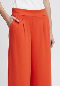 Oranje wijde broek met een gladde textuur, met een hoge tailleband en zijzakken. Inclusief voorplooien voor extra detail.