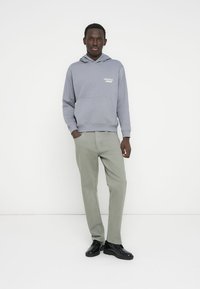 Γκρι φούτερ με κουκούλα, μπροστινή τσέπη και λευκά γράμματα, συνδυασμένο με ανοιχτοπράσινο παντελόνι και μαύρα παπούτσια. Μονή φιγούρα σε λευκό φόντο.