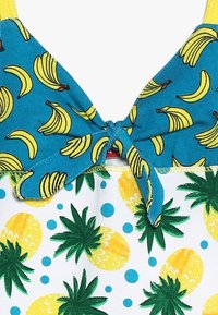 Top da bagno in tessuto turchese con stampe di banane gialle e dettaglio con fiocco, abbinato a una base bianca con ananas verdi e punti blu.
