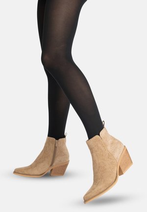 Jambes portant des collants noirs et des bottines en daim beige avec de petits talons carrés en bois et des fermetures éclair latérales, sur un fond uni clair.
