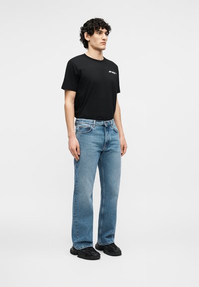 Homme aux cheveux bouclés portant un t-shirt noir, un jean bleu clair et des baskets noires, debout devant un fond blanc.