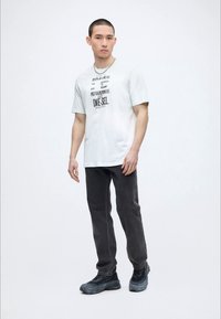 Νέος άνδρας με λευκό T-shirt με γραφικό, σκούρα τζιν και μαύρα αθλητικά παπούτσια, στέκεται μπροστά σε έναν απλό φωτεινό φόντο.