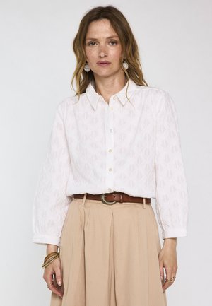 Femme portant un chemisier blanc texturé à boutons, une jupe plissée beige, une ceinture marron, des bracelets dorés et des boucles d'oreilles pendantes sur un fond uni.