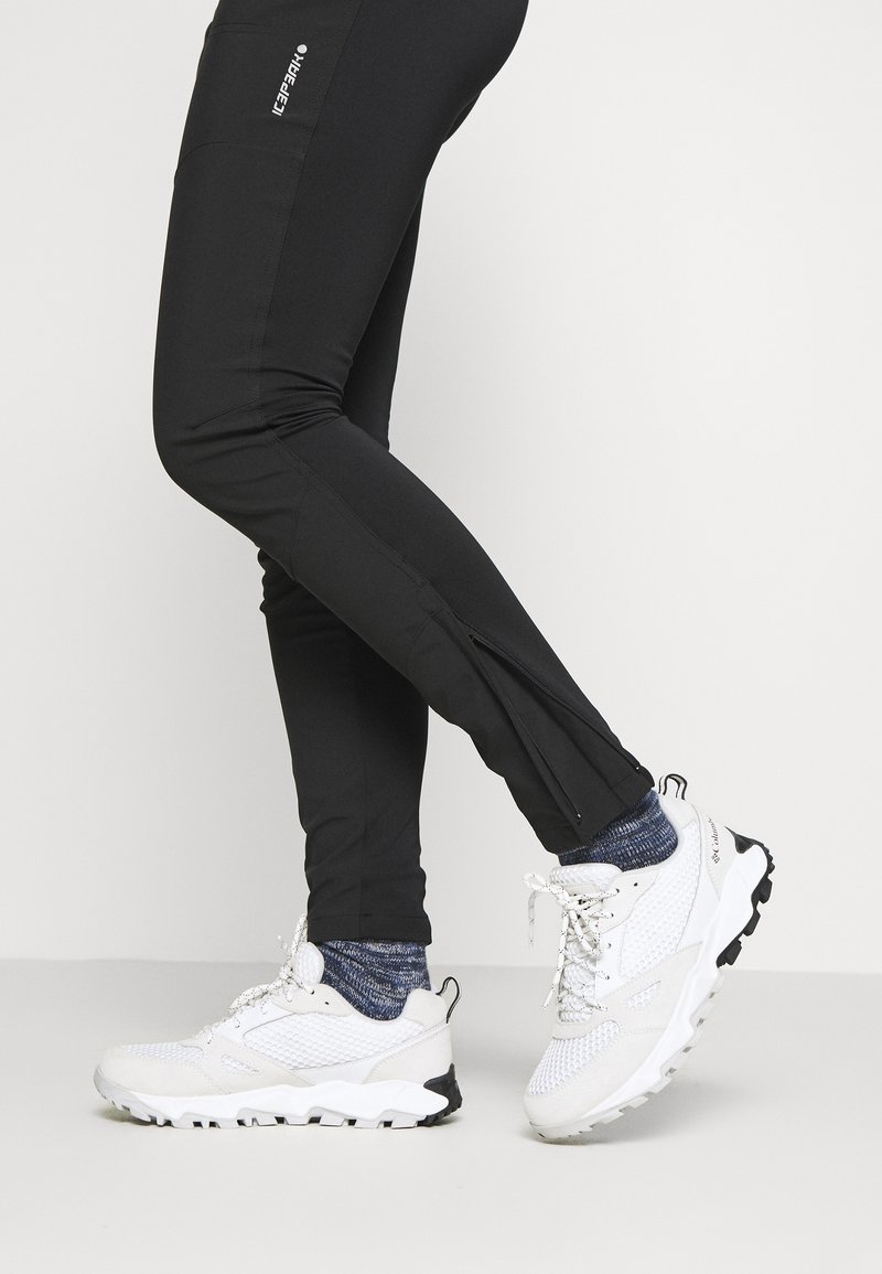 Pantaloni neri attillati con zip laterali, abbinati a sneakers bianche in mesh e gomma. I calzini sono blu navy con un motivo di texture.