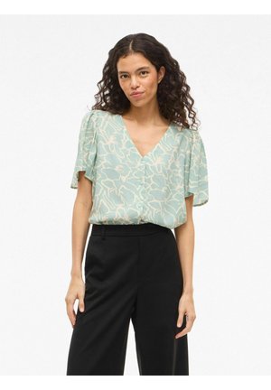 Kobieta z kręconymi włosami ubrana w miętową bluzkę w kwiaty z dekoltem w serek i czarne szerokie spodnie, stojąca na białym tle.