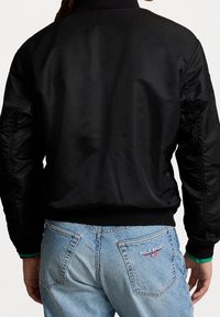 Svart bomberjacka med ribbat krage och manschetter. Slät, glänsande tyg med elastisk nederkant. Buren över ljusblå jeans med märkesdetalj.