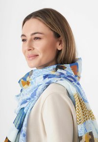 Écharpe multicolore avec des motifs bleus, dorés et bruns, fabriquée en tissu léger ; drapée lâchement autour du cou sur un haut clair.