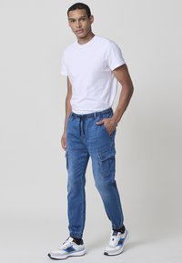 Lahka bela kratka majica s kratkimi rokavi v kombinaciji z modrimi denim cargo hlačami z elastičnimi manšetami, več žepi in pasom na vrvico.