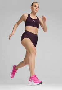 Conjunto deportivo de dos piezas en tela morada oscura; incluye un sujetador deportivo ajustado y un pantalón corto de tiro alto. Zapatillas de correr rosas con un diseño texturizado.