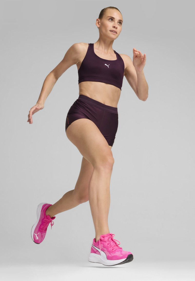 Conjunto deportivo de dos piezas en tela morada oscura; incluye un sujetador deportivo ajustado y un pantalón corto de tiro alto. Zapatillas de correr rosas con un diseño texturizado.