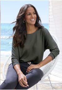 Frau mit langen dunklen Haaren, die ein grünes Langarmshirt und dunkle Hosen trägt, sitzt auf einem weißen Stuhl am Meer, lächelt und schaut nach rechts.