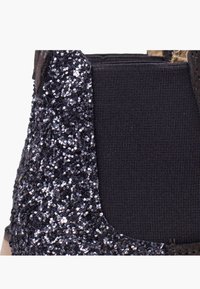 Primer plano de un zapato de tela oscura con detalle de purpurina negra y panel elástico negro en el costado.