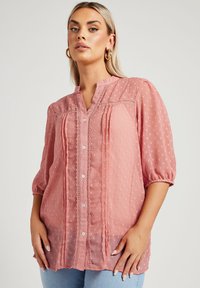 Blusa rosa con un pattern a pois testurizzati, con bottoni frontali, maniche a sbuffo corte e dettagli in pizzo lungo lo scollo e il davanti.