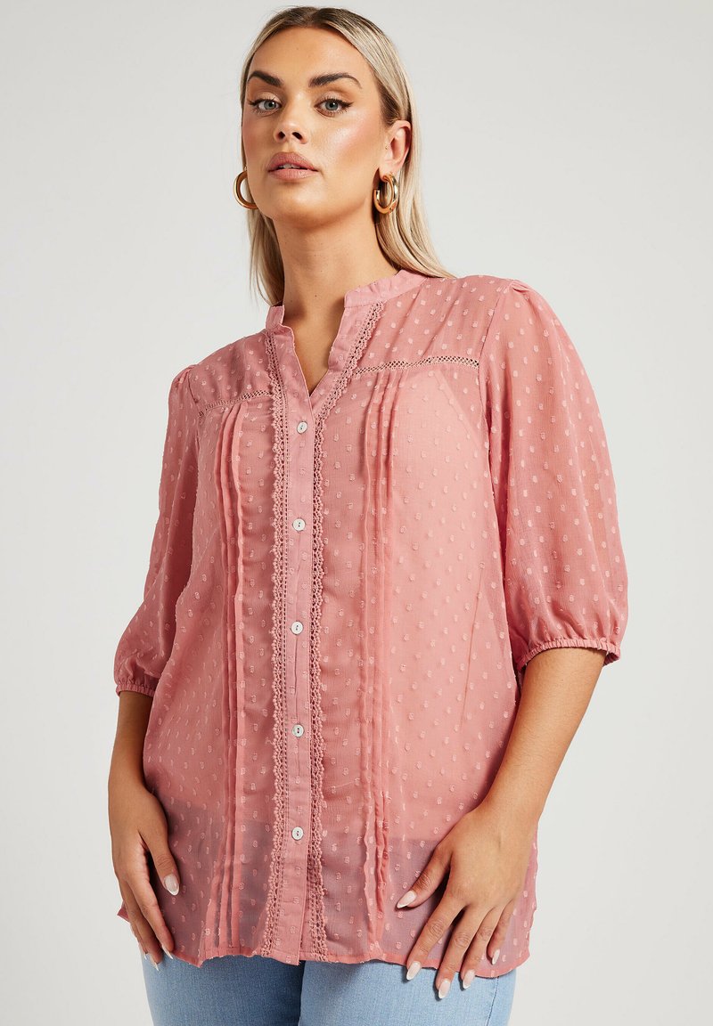 Blusa rosa con un pattern a pois testurizzati, con bottoni frontali, maniche a sbuffo corte e dettagli in pizzo lungo lo scollo e il davanti.