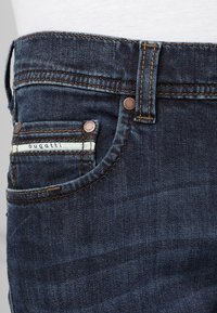 Jean oscuro de mezclilla con un diseño clásico de cinco bolsillos, costuras en marrón contrastante y una pequeña etiqueta con el logo en el bolsillo delantero.