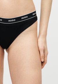 Gros plan d'une personne portant un slip noir de la marque HUGO, montrant la ceinture et une partie de la cuisse et de la main.