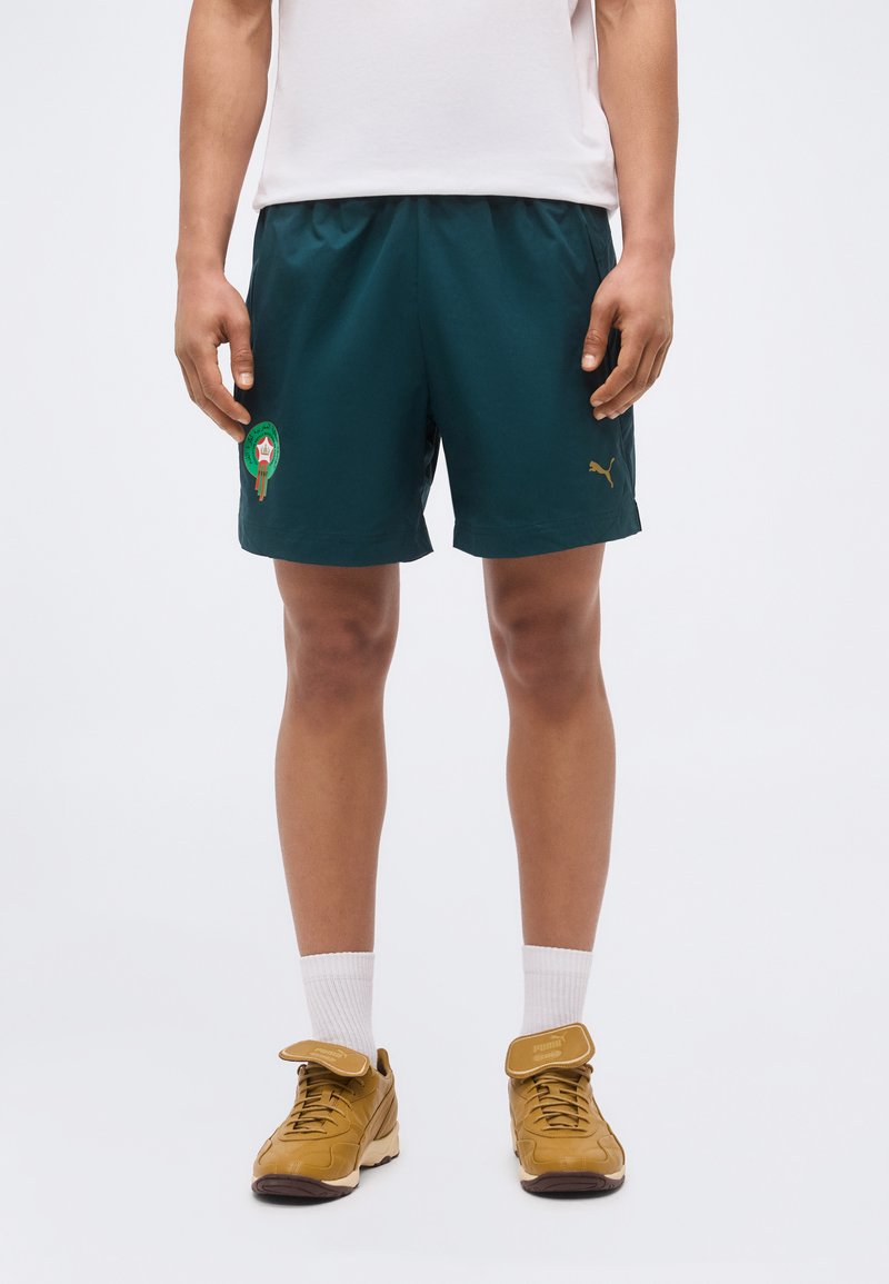 Άτομο που φοράει σκούρα πράσινα σορτς Puma με λογότυπο στον αριστερό μηρό, λευκές κάλτσες και μπεζ παπούτσια Puma με τη γλώσσα λυγισμένη προς τα εμπρός.