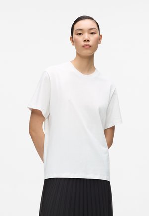 Mlada osoba sa glatkom kosom nosi jednostavnu bijelu majicu kratkih rukava i crnu plisiranu suknju, stojeći na svijetloj pozadini.