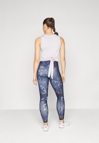 Ljust lila ärmlös topp med detalj av knytning bak, tillsammans med mörka mönstrade leggings med blå och vita marmoreringar. Vita sneakers.