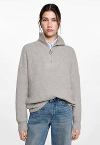 Hellgrauer, gerippter Strickpullover mit hohem Kragen, Reißverschluss und Ringgriff. Getragen über einem hellblauen Hemd mit Knöpfen, kombiniert mit Denim.