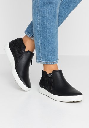 Osoba nosząca czarne skórzane i materiałowe sneakersy za kostkę z białymi podeszwami i bocznymi zamkami błyskawicznymi, w połączeniu z jasnoniebieskimi dżinsami.