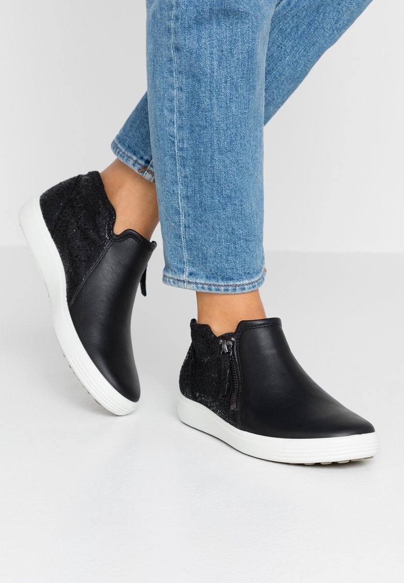 Personne portant des baskets montantes en cuir et tissu noir avec des semelles blanches et des fermetures éclair latérales, associées à un jean bleu clair.