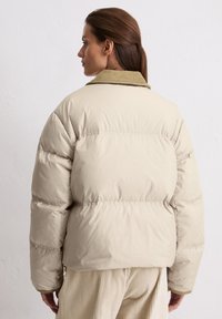 Jachetă puffer bej cu guler din catifea, având un design matlasat, mansete elastice și o croială relaxată, purtată peste pantaloni bej texturați.