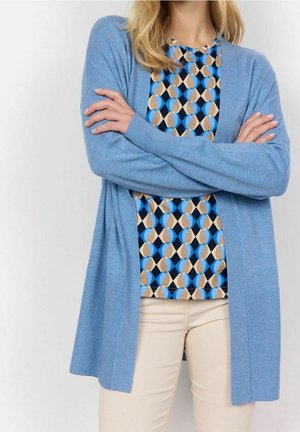 Femme les bras croisés, portant un cardigan bleu par-dessus un haut à motif géométrique et un pantalon beige, sur un fond uni.