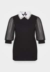 Svart topp med en vit krage som har en dekorativ pärldetalj, genomskinliga puffärmar med prikar och en figurnära passform.