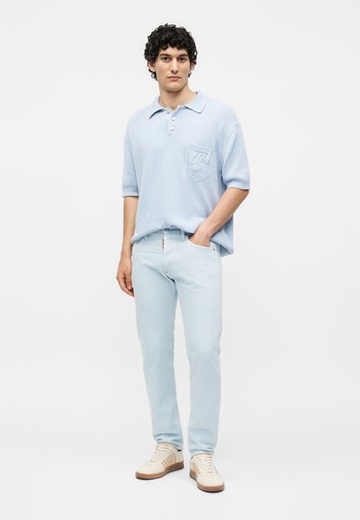 Άνδρας που φοράει ανοιχτό μπλε πόλο με κοντά μανίκια, ανοιχτό μπλε τζιν και λευκά αθλητικά με καφέ σόλες, στέκεται με το ένα χέρι στην τσέπη.