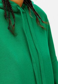Sudadera verde con una superficie texturizada, que cuenta con una capucha con cordones y mangas cortas, enfocándose en el color vibrante y el ajuste relajado.