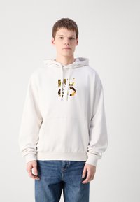 Sudadera blanca con un bolsillo canguro, que presenta un gran logo en oro y negro en el pecho. Los puños y el dobladillo son acanalados. Se llevan unos vaqueros abajo.