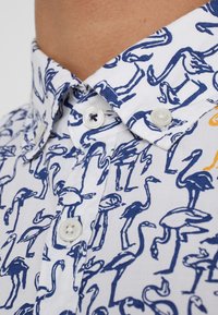 Camicia bianca a maniche corte con un motivo di fenicotteri blu, con colletto button-down e dettagli sui bottoni in contrasto.