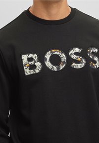 Svart sweatshirt med en texturerad "BOSS"-logotyp i vitt och orange, med rund hals och långa ärmar. Slät tygtextur.