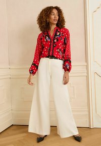 Sarkans rakstu blūze ar punktiņiem, melniem akcentiem un pogājamu priekšu, kombinēta ar baltām platiem biksēm un melniem šauriem apaviem.