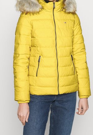 Kobieta w żółtej pikowanej kurtce z kieszeniami na zamek, kapturem obszytym futerkiem i niebieskich dżinsach widocznych od połowy ud do szyi.