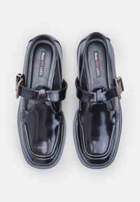 Un par de brillantes zapatos de charol negro tipo Mary Jane con punteras cuadradas y correas de hebilla dorada, etiquetados como Bata Red Label en su interior.
