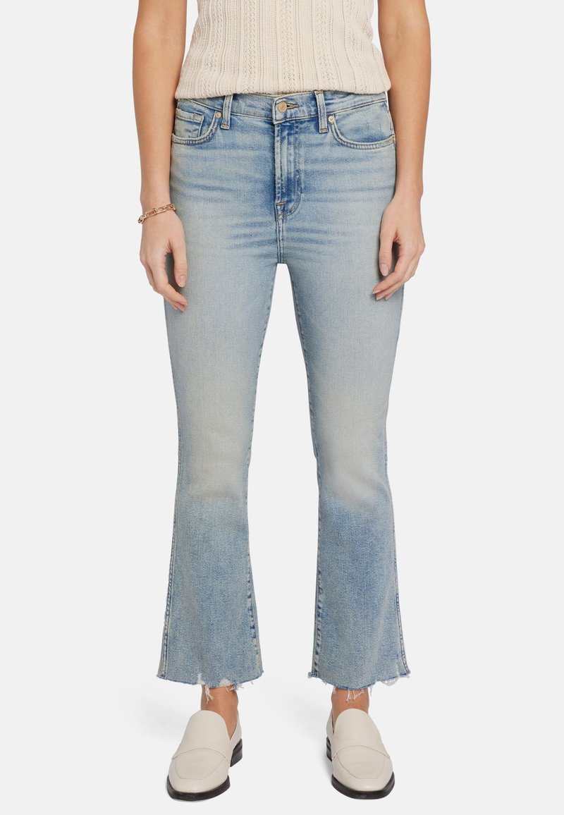 Jean en denim bleu clair, évasé, avec taille haute, finition délavée et ourlet brut. Associé à des chaussures à enfiler de couleur claire.