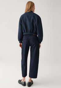 Giacca bomber navy con orlo arricciato, abbinata a jeans scuri a gamba dritta e scarpe nere slip-on. Dettagli di cucitura sottili visibili.