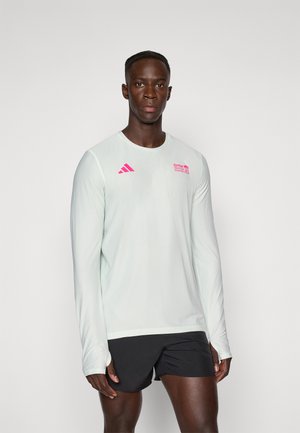 Atleta masculino con una camiseta blanca de manga larga con logotipos rosas y pantalones cortos de correr negros, de pie contra un fondo claro y sencillo.