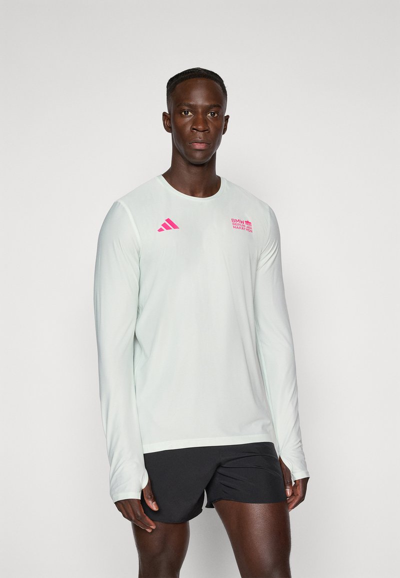 Männlicher Sportler in einem weißen Langarmshirt mit rosa Logos und schwarzen Laufshorts, der vor einem schlichten hellen Hintergrund steht.