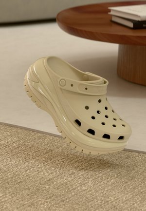 Sabot beige à enfiler avec des trous de ventilation et une sangle, flottant au-dessus d'un tapis beige texturé près d'une table ronde en bois avec des livres.