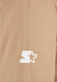 Beige színű pamut póló, bal oldalon fehér grafikával, amelyen egy "S" és csillag minta található, sima textúrával és egyszerű kivitelezéssel.