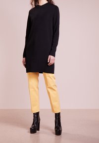 Abito lungo nero a maniche lunghe con scollo rotondo, abbinato a pantaloni gialli aderenti e stivali alla caviglia neri con platform. Tessuti lisci, design minimale.