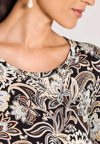 Vrouw met parel-druppelsieraden en een zwart bovenstuk met een ingewikkeld beige, witte en bruine bloemige paisley-patroon, waarbij de nek en schouderdetails zichtbaar zijn.